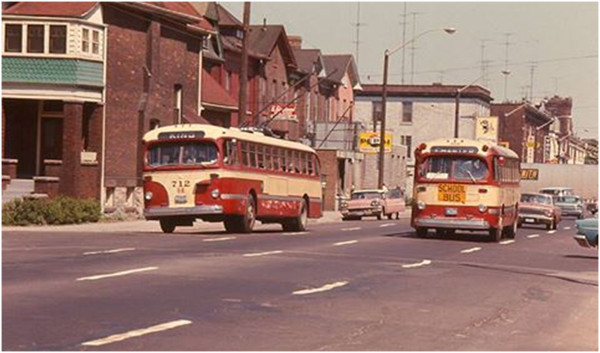 1950s era maroon and cream Hamilton Street Railway trolley buses crossing West Ave on King. St John steeple is at upper right.